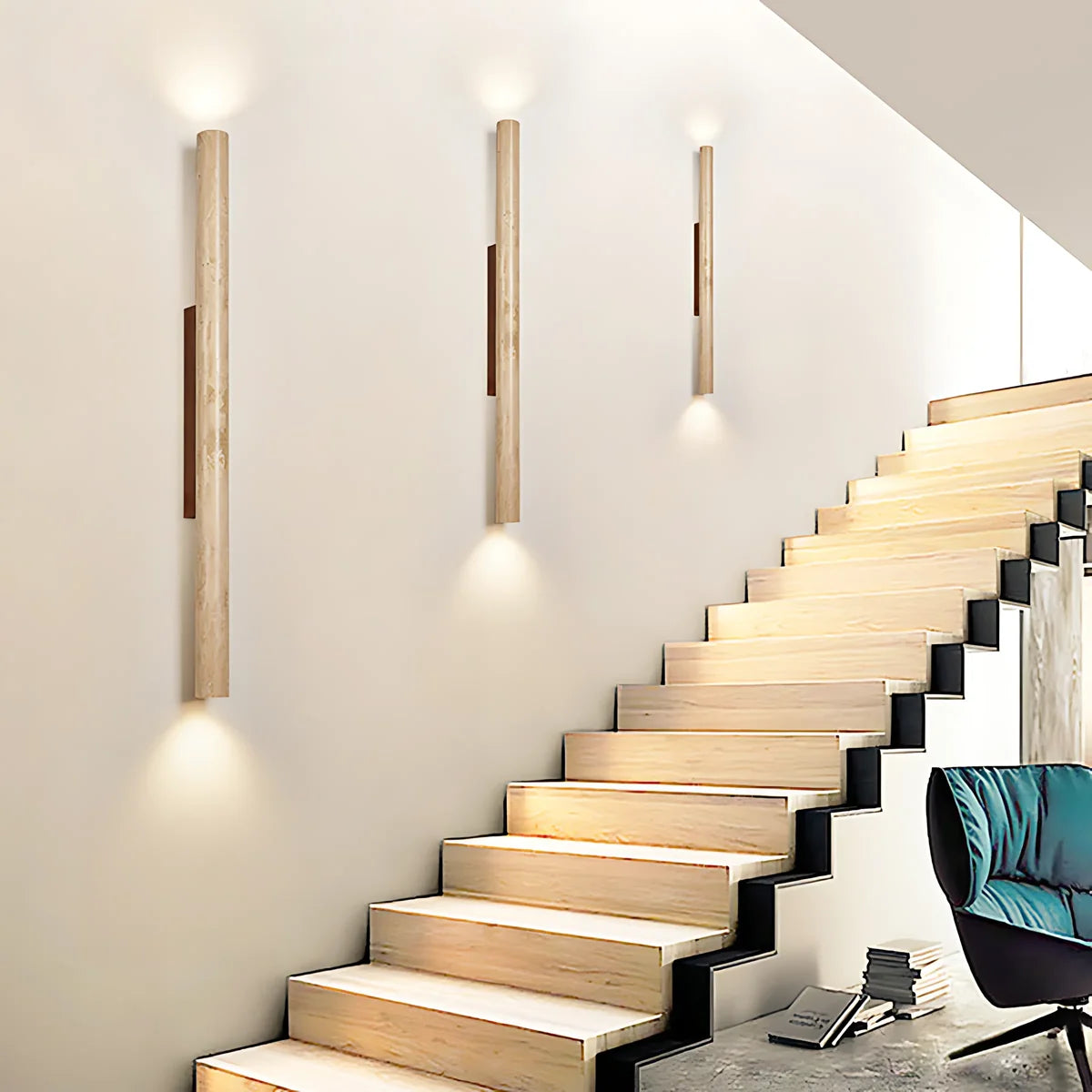Three modern wall sconces with vertical light wood posts and dark center panels are mounted on a cream wall above a wooden staircase. A teal chair is partially visible in the lower right corner.