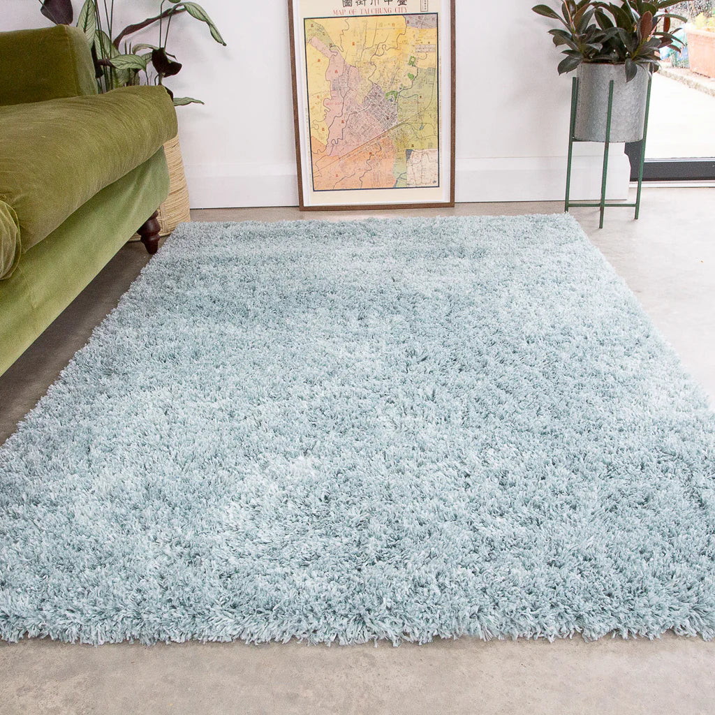 A light blue shag area rug laid on a concrete floor in a modern living room setting, with a green sofa on the left and a framed map on the wall behind.