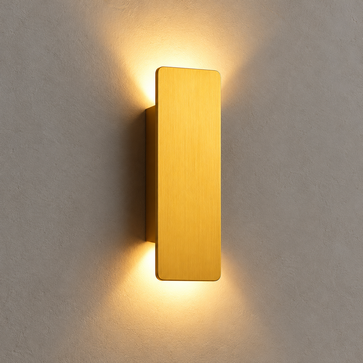 A modern rectangular wall sconce with a brushed gold finish, mounted on a textured gray wall and casting a warm yellow light downward and upward.