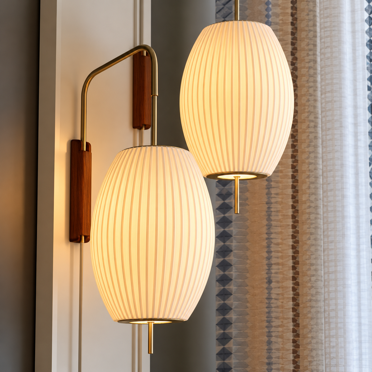 Two modern wall-mounted sconces with ribbed white cylindrical shades and warm brass arms, shown lit in an interior setting beside patterned curtains.