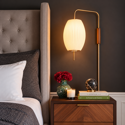 A mid-century modern floor lamp with a ribbed white pleated shade and a curved brass arm, illuminated in a bedroom setting next to a bed and wooden nightstand.