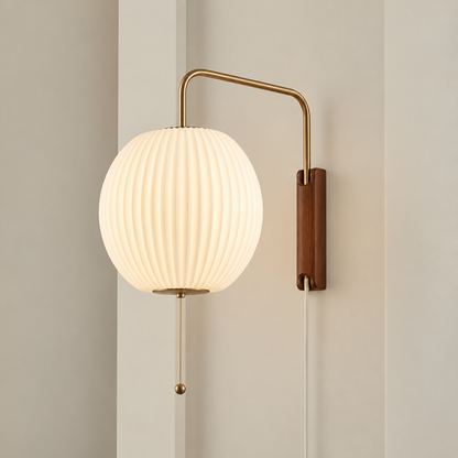 A modern wall-mounted sconce with a ribbed white globe shade, a curved brass metal arm, a vertical brown wood-look wall plate, and a hanging pull chain, shown lit against a beige wall.