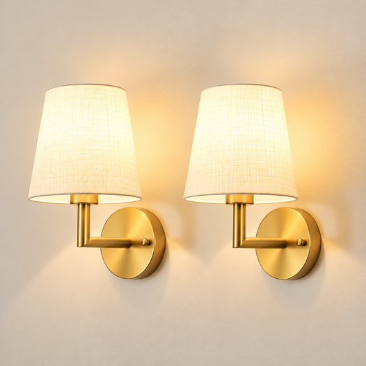 A pair of modern wall sconces with warm gold-finished metal bases and tapered off-white fabric shades, shown lit against a plain beige wall.