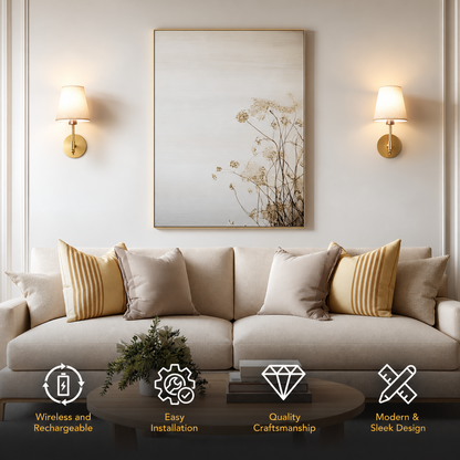 A beige fabric sofa in a styled living room setting, decorated with striped and solid beige pillows, a small potted plant on a dark wooden coffee table, and a large vertical framed botanical art print on the wall above. Two gold wall sconces are lit on either side of the artwork.