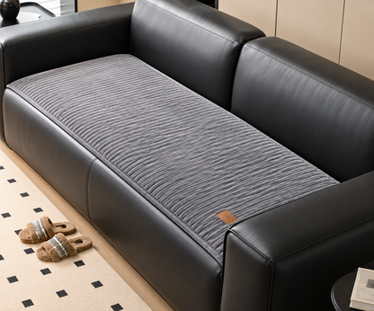 A black leather sofa with a ribbed gray cushion on the seat, shown in a styled interior with a patterned rug and slippers nearby.