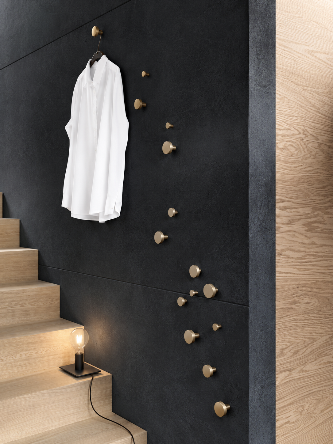 A white long-sleeve button-up shirt hangs from a brass wall hook on a dark charcoal textured wall beside a light wood staircase. Multiple matching brass wall hooks are mounted along the wall, with a small black floor lamp on the step below.