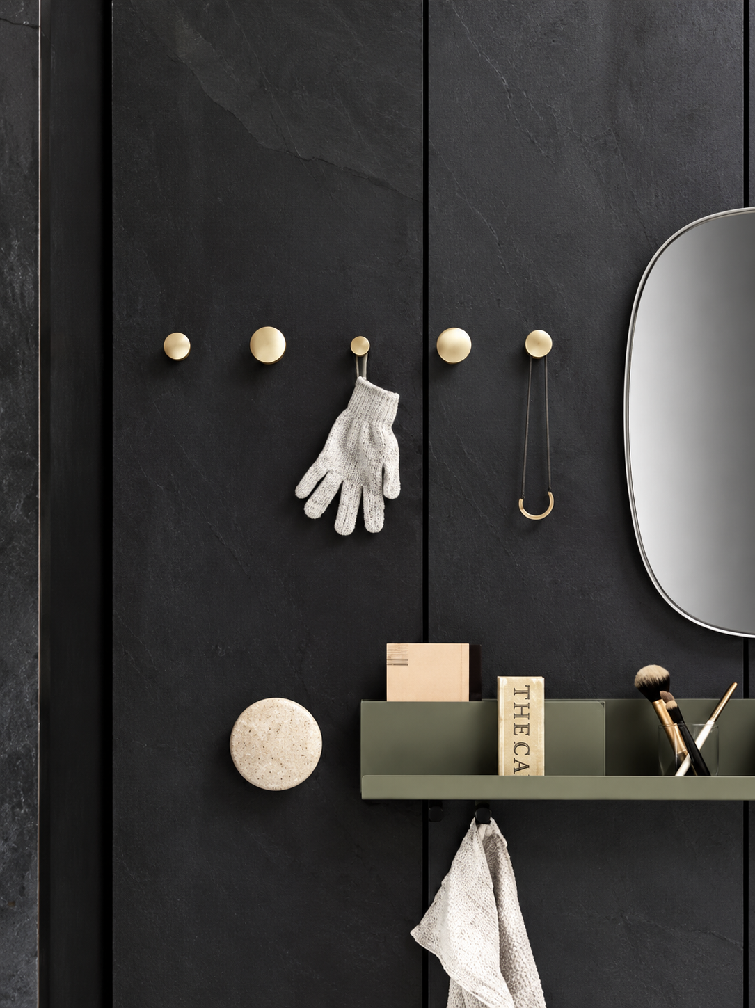 A styled bathroom or vanity scene with a dark charcoal wall featuring brass wall hooks, a white knit glove hanging on one hook, a green shelf with toiletries, and part of a round mirror visible on the right.