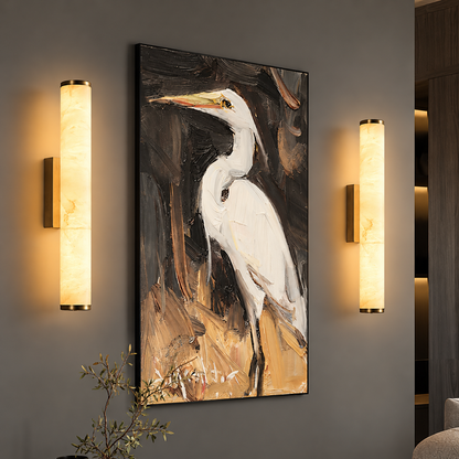 A vertical framed wall art print depicting a white heron on a dark, painterly background, displayed on a gray wall between two illuminated cylindrical wall sconces.