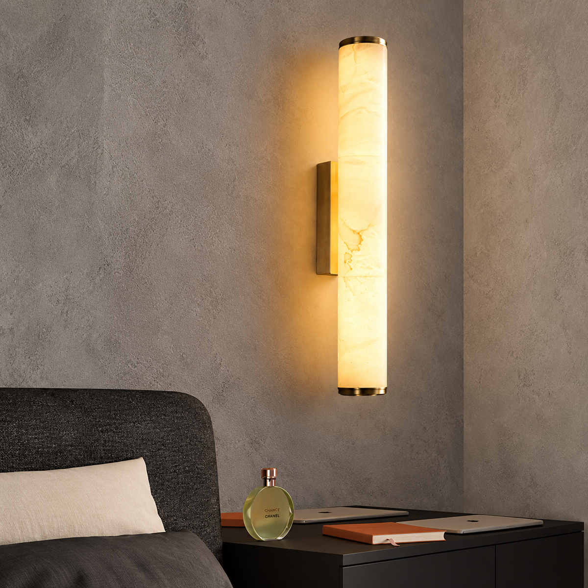 A modern wall sconce light fixture with a cylindrical illuminated white marble-look column and gold-toned metal accents, mounted on a textured gray wall above a dark bedside table.
