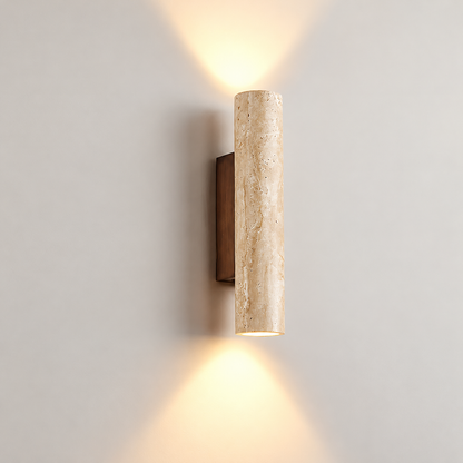 A modern cylindrical wall sconce with a textured beige travertine stone column and a dark bronze wall mount, emitting warm light upwards and downwards against a neutral gray wall.