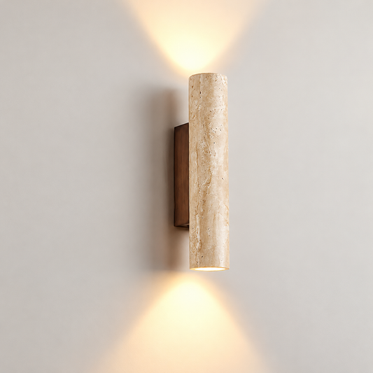 A modern cylindrical wall sconce with a textured beige travertine stone column and a dark bronze wall mount, emitting warm light upwards and downwards against a neutral gray wall.