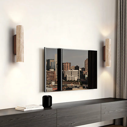 A modern wall-mounted TV with a mirrored finish reflecting a city skyline, flanked by two cylindrical wall sconces on a white wall above a dark wood console.