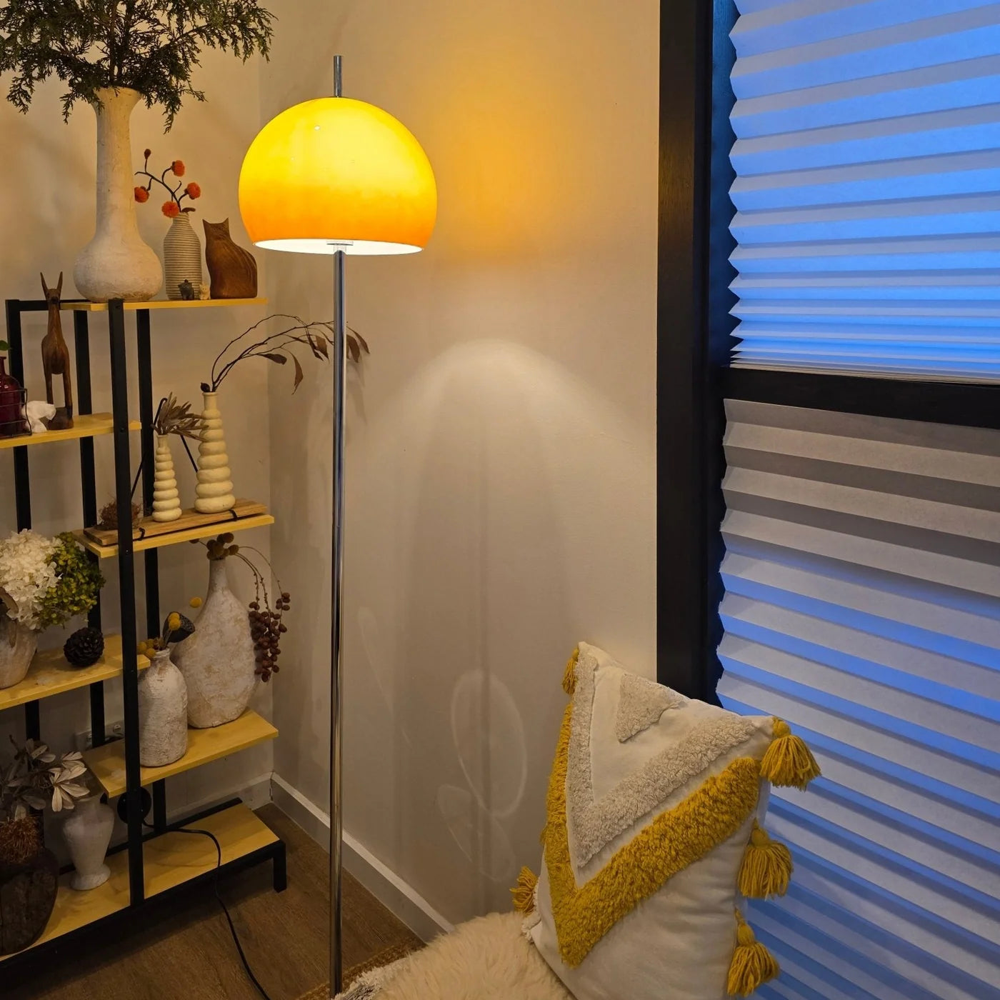 A modern floor lamp with a large, spherical, dome-shaped shade glowing warm yellow, standing in a styled room next to a window with blue-toned blinds and a decorative shelf.