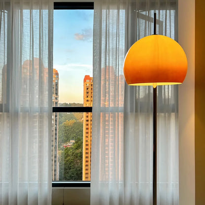 A modern floor lamp with a large, dome-shaped warm yellow shade stands in a room next to a window with sheer white curtains, looking out onto a city skyline at sunset.