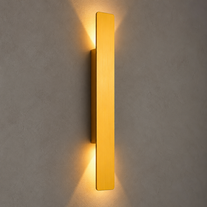 A modern vertical rectangular wall sconce in a brushed gold finish, emitting a warm yellow light against a textured gray wall.