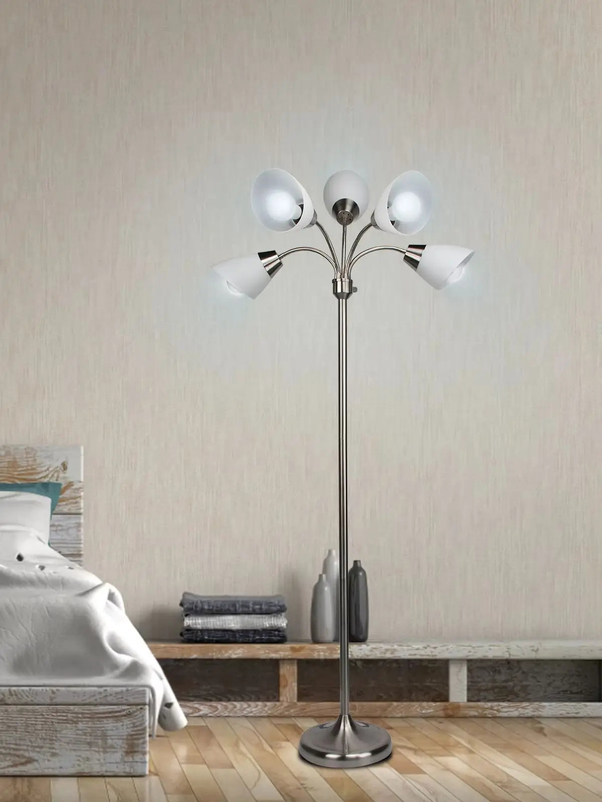 A modern multi-light floor lamp with a chrome-finished stand and five white glass shades, illuminated in a bedroom setting with a bed and wooden floor visible.