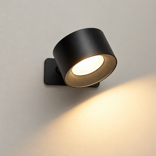A modern black cylindrical wall sconce light fixture mounted on a neutral gray wall, casting a warm yellow glow downwards and to the right.