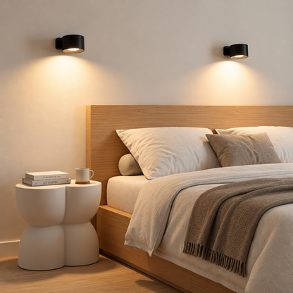 A modern bedroom scene with a wooden bed frame, white bedding, a brown throw blanket, a sculptural white side table with a mug, and two black wall sconces mounted on the wall above the bed.