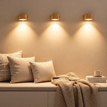 Three cylindrical wall sconces with warm wood-look shades mounted on a beige wall above a cushioned bench with pillows and a throw blanket, casting soft warm light downward.