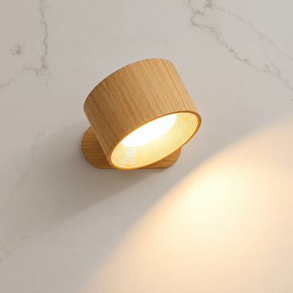 A modern cylindrical wall sconce with a light natural wood grain finish, shown illuminated against a white marble-textured wall.