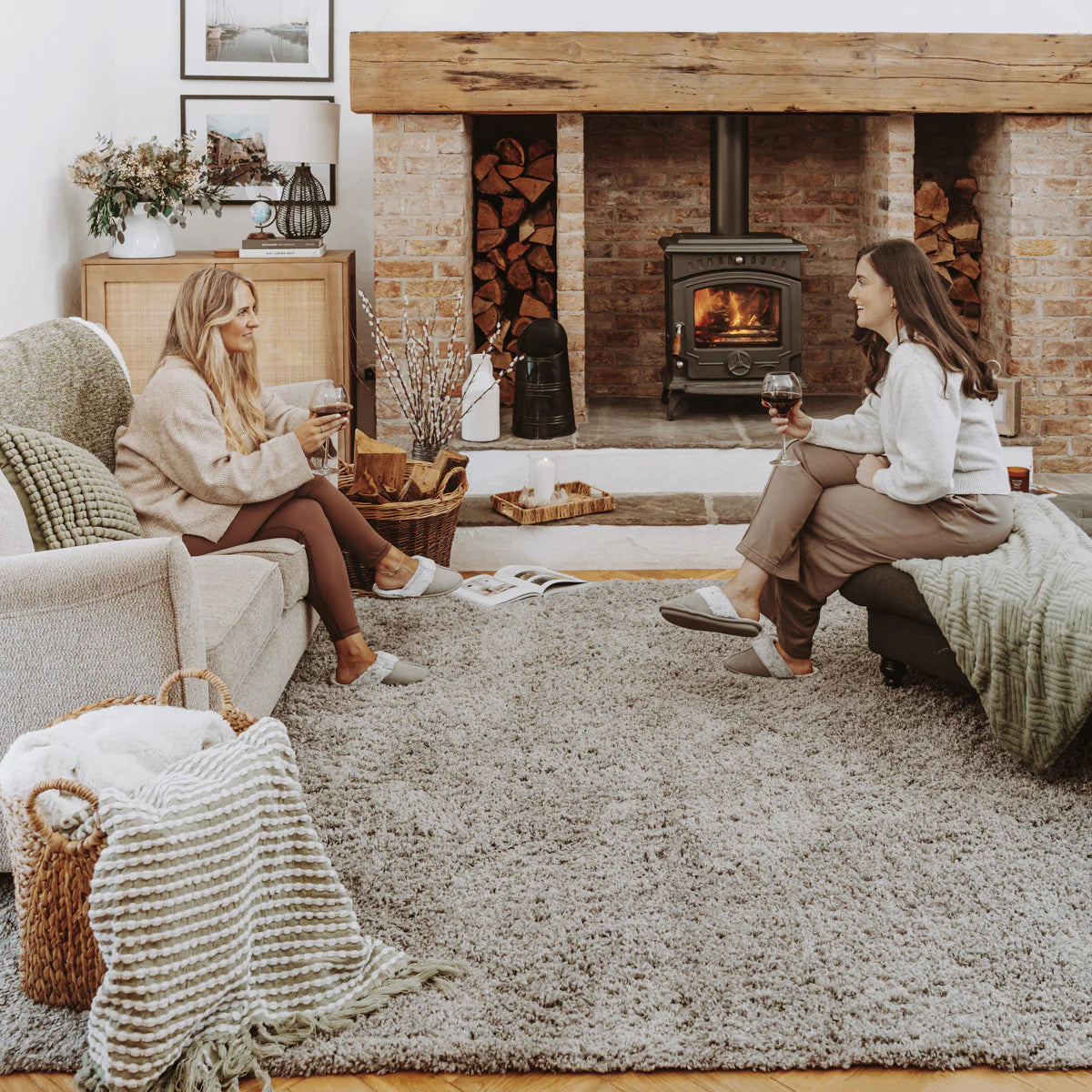 A large, plush light gray shag area rug covers the floor in a cozy living room setting, with two women sitting on upholstered chairs on either side of it, engaged in conversation near a brick wood-burning stove.