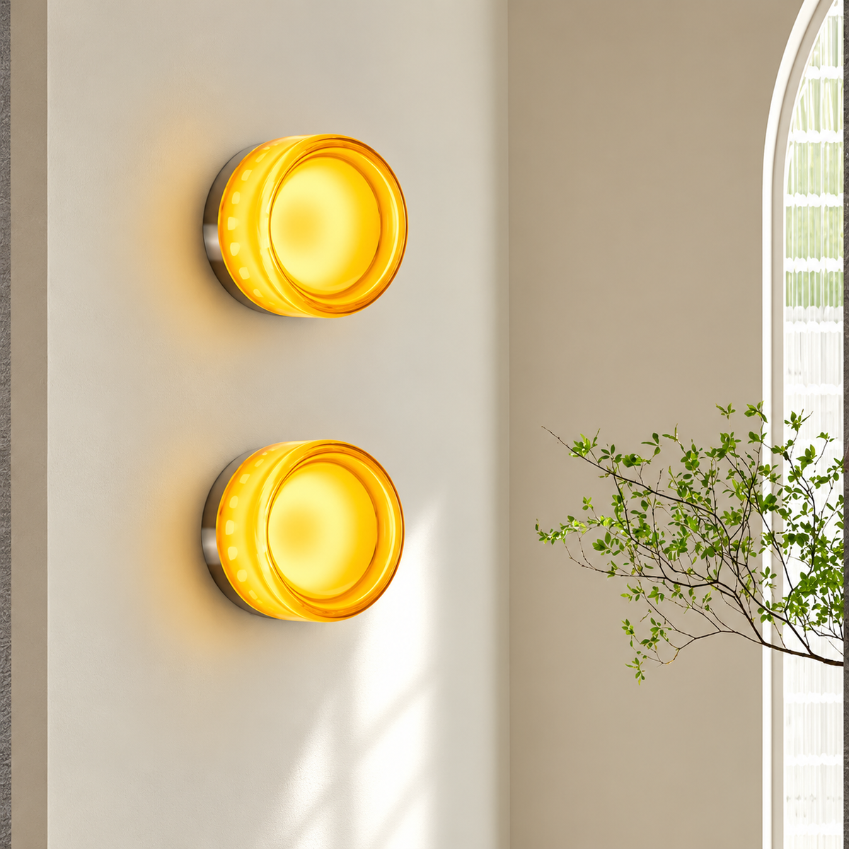 Two modern wall sconces with glowing amber-yellow circular glass shades mounted vertically on a beige wall, with a leafy branch visible at the right edge.