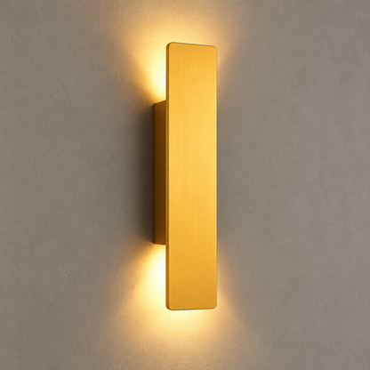 A modern vertical rectangular wall sconce in brushed gold, mounted on a textured gray wall and casting a warm yellow light downward and upward.