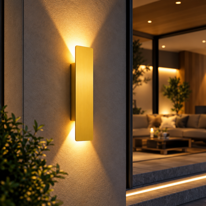 A modern vertical rectangular wall sconce in a warm brushed gold finish, mounted on a textured beige exterior wall and casting a soft golden glow. The background shows a softly blurred contemporary living room interior with warm ambient lighting.