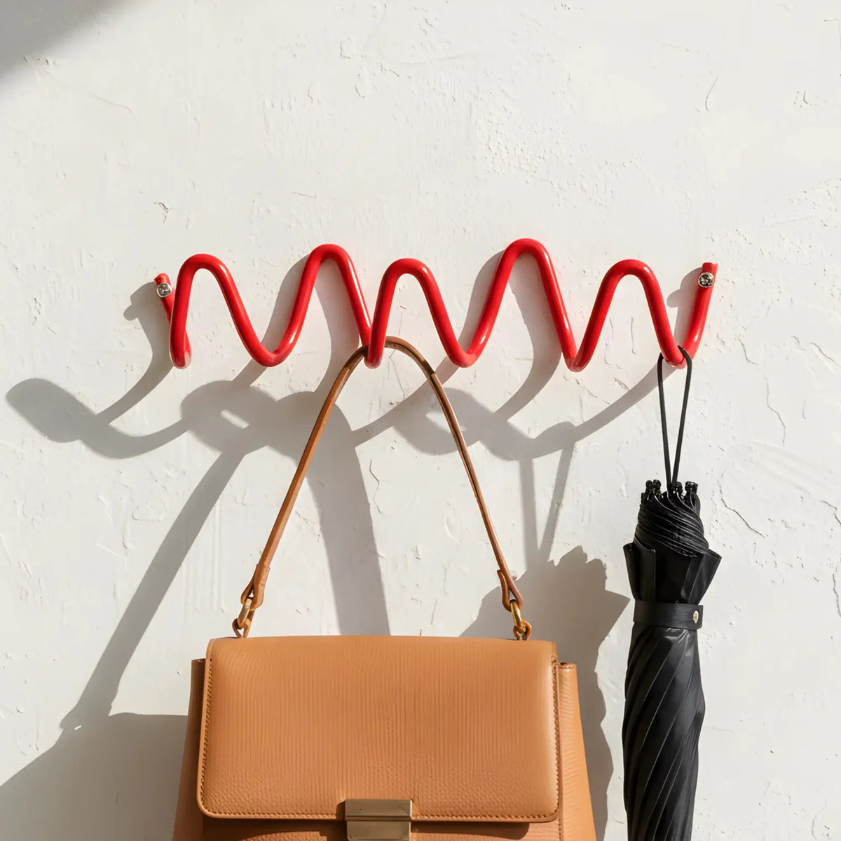 A bright red wavy metal wall hook rack mounted on a textured white wall, holding a tan leather handbag and a black umbrella.