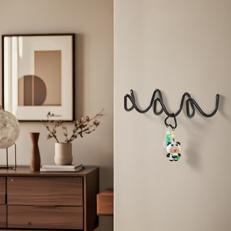 A black wavy spiral wall hook mounted on a beige wall, holding a small cartoon keychain, with a styled interior scene on the left including a wooden cabinet, framed art, and vases.
