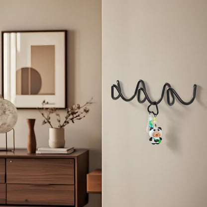 A black wavy spiral wall hook mounted on a beige wall, holding a small cartoon keychain, with a styled interior scene on the left including a wooden cabinet, framed art, and vases.