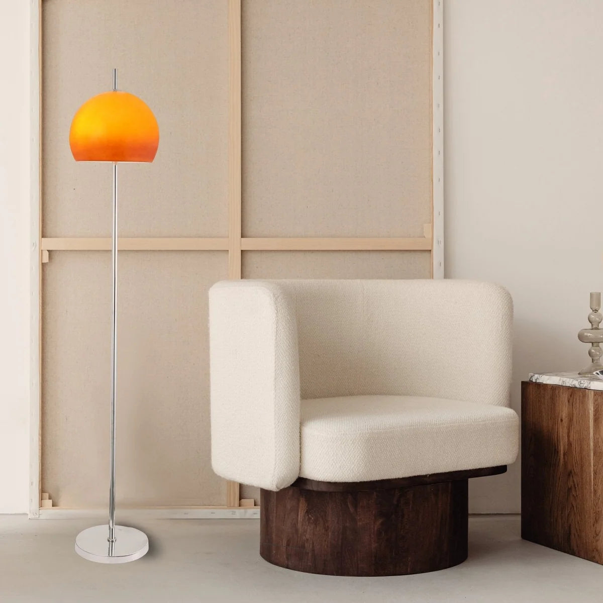 A modern floor lamp with a glowing orange dome shade and a slim silver stand, positioned next to a cream-colored upholstered armchair with a dark wood base in a minimalist room setting.